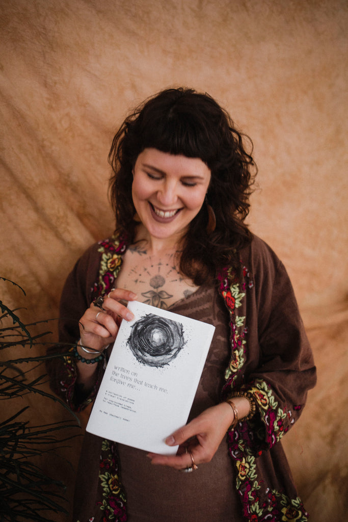 Rachael Amber smiles holding her cyclical poetry book "written on the trees that teach me, forgive me..."